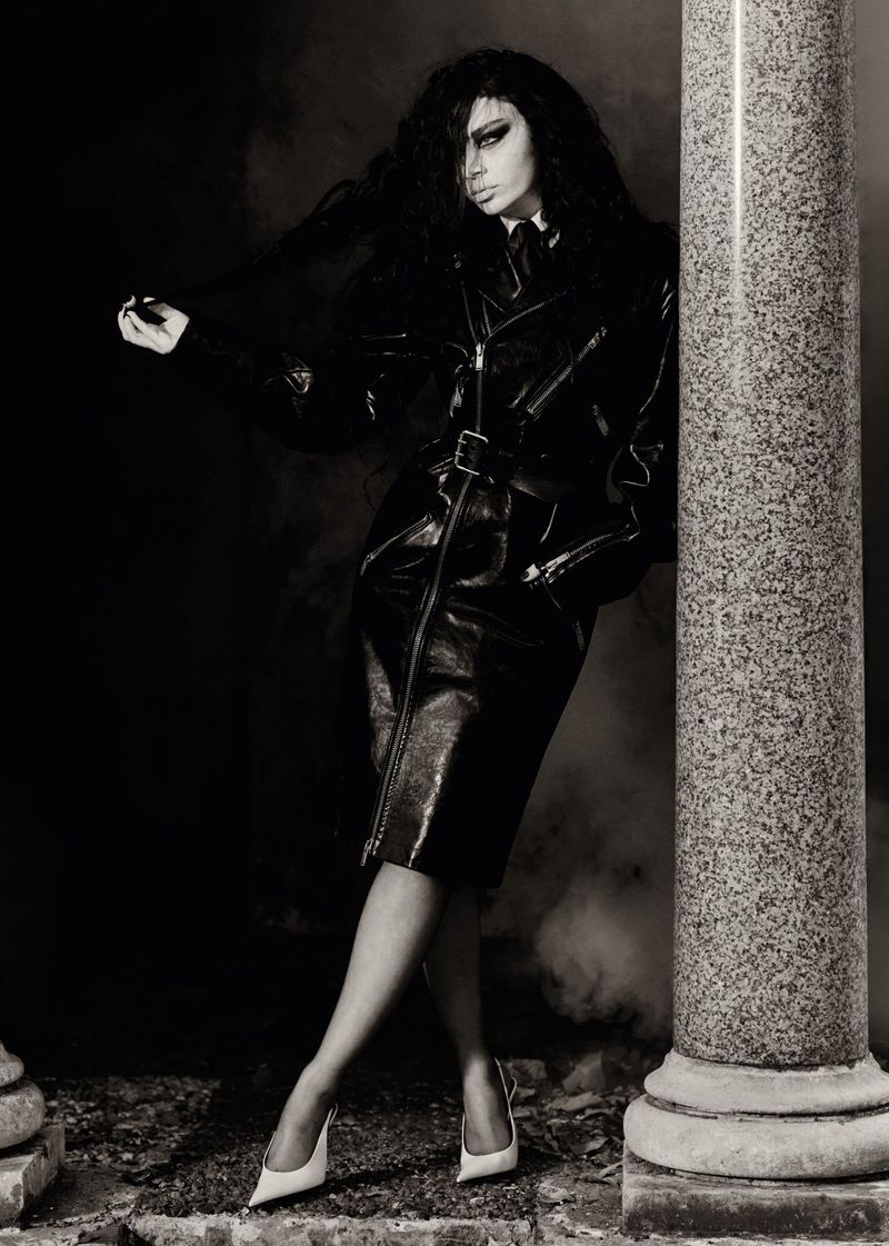 Fashionable adult female posing in a black leather skirt and jacket, leaning against a stone pillar, in a monochromatic outdoor setting.