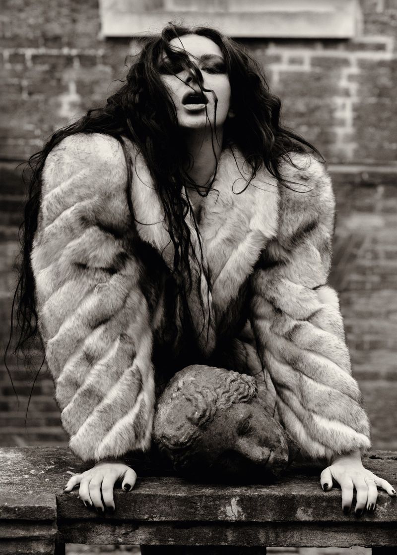 Black and white portrait of a woman with long dark hair wearing a fur coat and resting her hands on a stone bust outdoors.