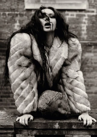 Black and white portrait of a woman with long dark hair wearing a fur coat and resting her hands on a stone bust outdoors.
