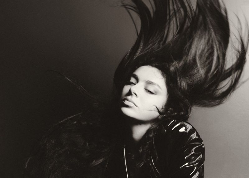 Monochromatic portrait of a woman with flowing hair wearing a shiny black jacket, eyes closed, set against a dark gray background.