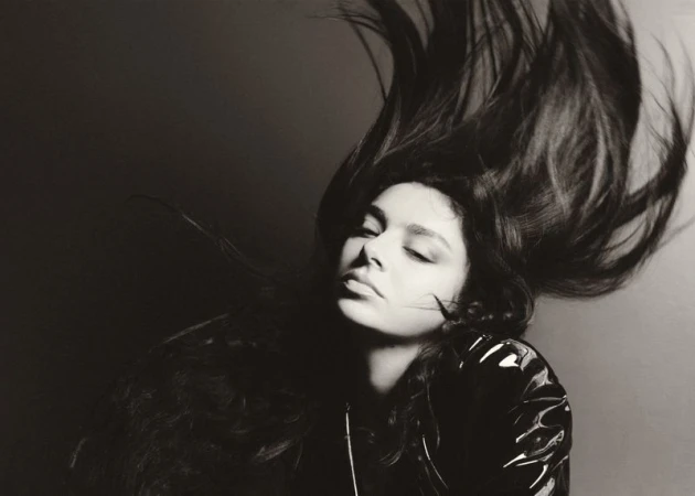 Monochromatic portrait of a woman with flowing hair wearing a shiny black jacket, eyes closed, set against a dark gray background.