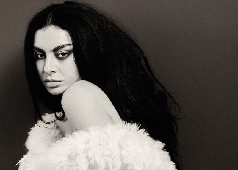 Monochromatic portrait of a woman with dark hair and makeup, draped in a white, furry garment against a dark grey backdrop.