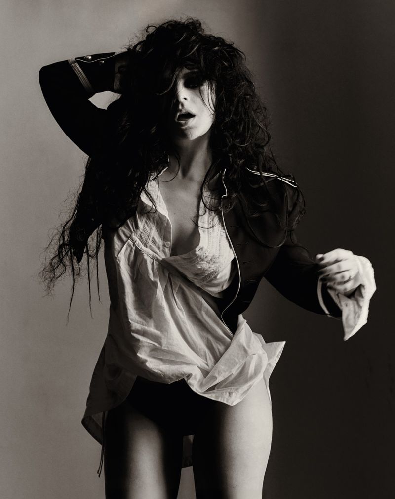 Monochrome portrait of a woman with shaggy dark hair posing dynamically in an open white shirt and dark military jacket.