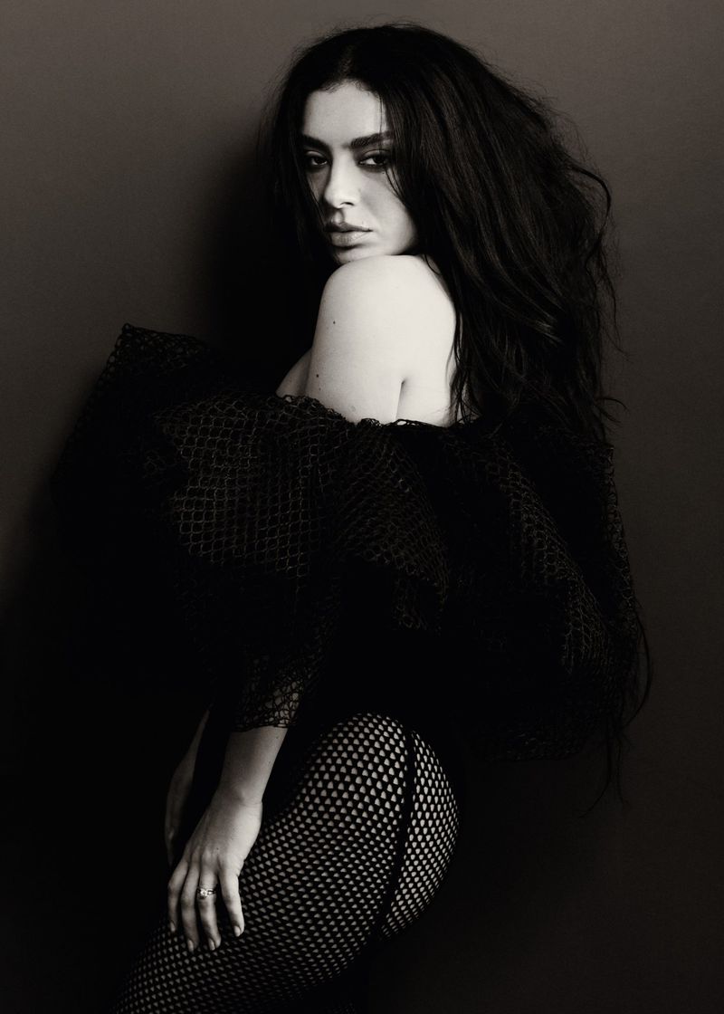 Monochrome portrait of a woman with dark hair, posing in a black net garment against a dark backdrop, looking over her shoulder.