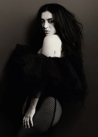 Monochrome portrait of a woman with dark hair, posing in a black net garment against a dark backdrop, looking over her shoulder.