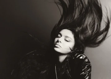 Model with long dark hair whipping through the air in a black glossy jacket against a neutral backdrop.