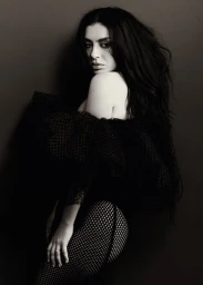 Charli XCX posing in an off-the-shoulder black mesh top and fishnet tights in a monochrome portrait against a dark backdrop.