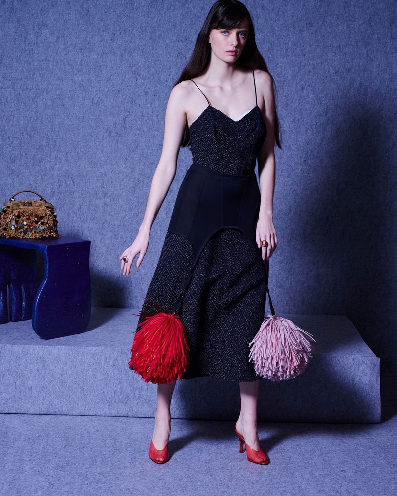 A woman poses in a black spaghetti-strap dress with red and pink pom-pom bags against a grey studio backdrop on a blue stool.