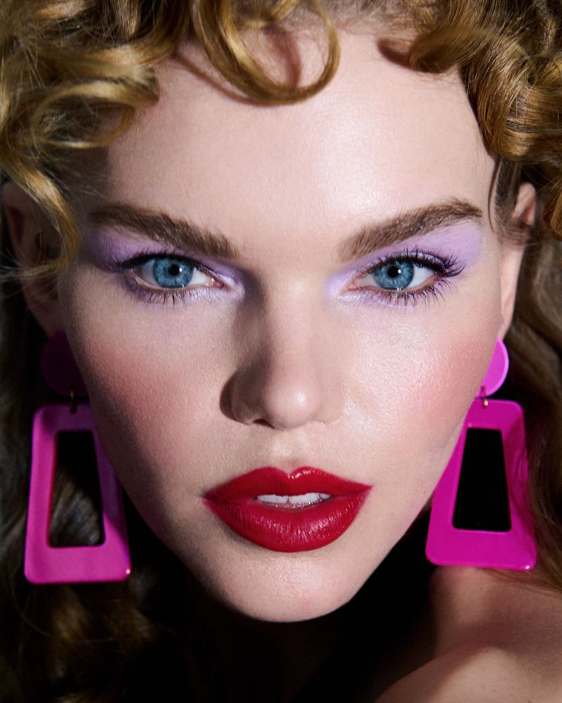Close-up shot of a woman with curly blonde hair, fair skin, blue eyes, red lipstick, and purple eyeshadow wearing pink earrings.