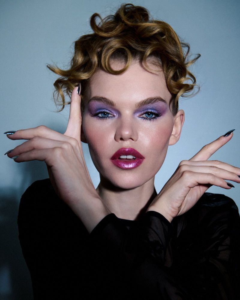 Close-up portrait of a woman with curly blonde hair and lavender makeup posing with her hands, set against a light blue backdrop.