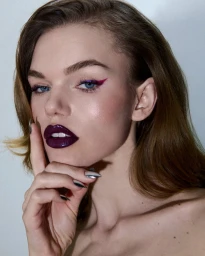 Model Daphne Maas posing with glossy dark plum lipstick, winged purple eyeliner, and metallic silver nails against a white background.