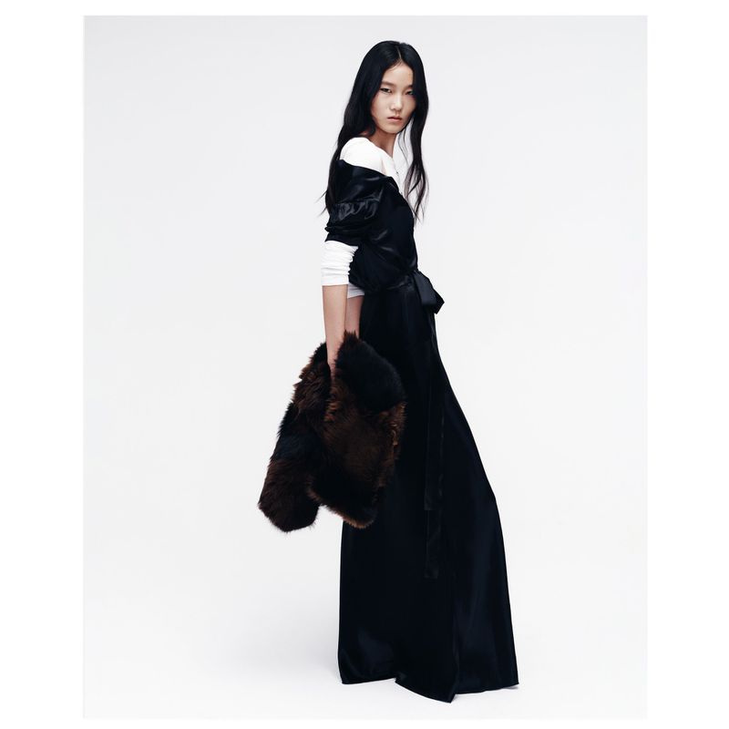 An Asian fashion model poses indoors, wearing a black dress and holding a dark fur jacket against a clean, light background.