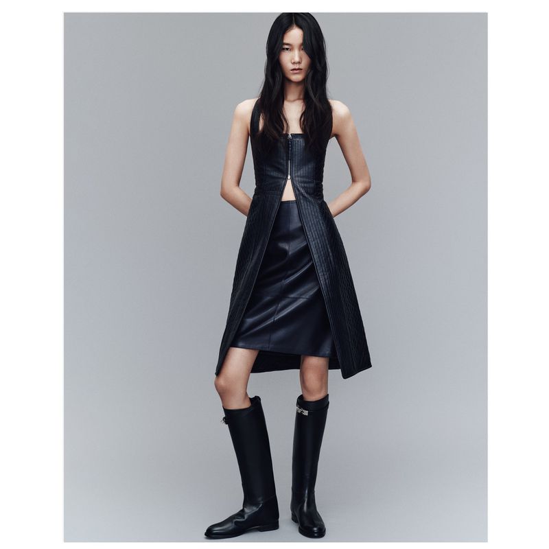 An Asian woman stands in a gray studio wearing a black, strapless leather dress and black boots, presenting a high-fashion editorial look.