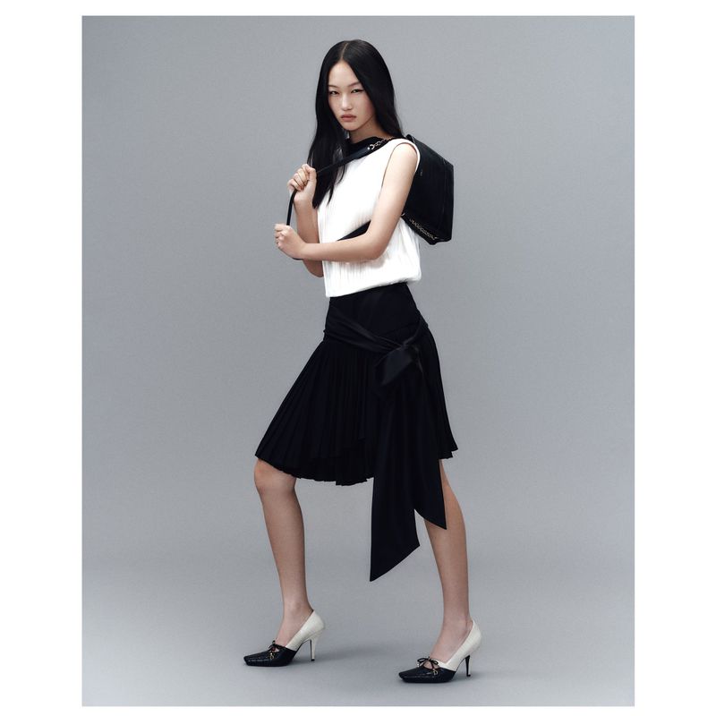 Full-length shot of a stylish Asian woman in a white top and black skirt, carrying a black bag, posing against a grey backdrop.