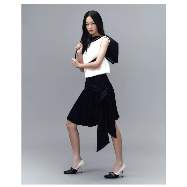 Full-length shot of a stylish Asian woman in a white top and black skirt, carrying a black bag, posing against a grey backdrop.