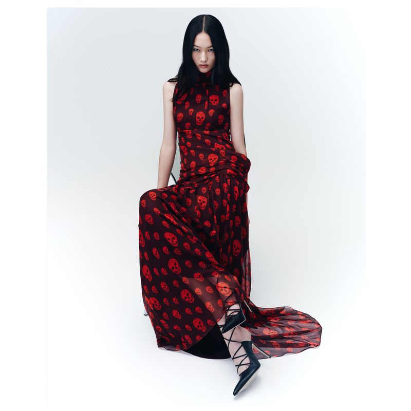 A slender woman with dark hair poses in a skull-print, sleeveless maxi dress with black lace-up heels in a brightly lit studio setting.