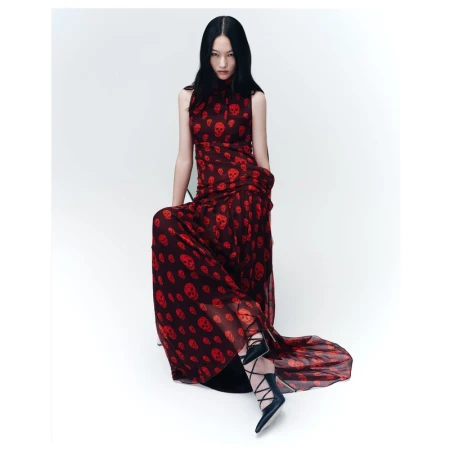 A slender woman with dark hair poses in a skull-print, sleeveless maxi dress with black lace-up heels in a brightly lit studio setting.