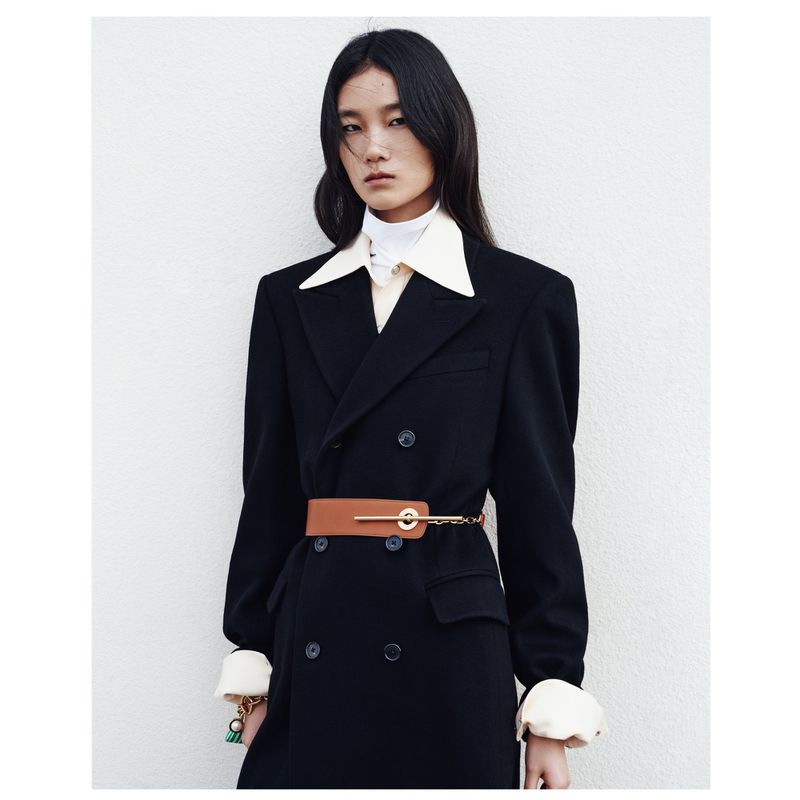 A stylish Asian woman poses indoors, showcasing a double-breasted black coat cinched with a tan belt against a neutral backdrop.