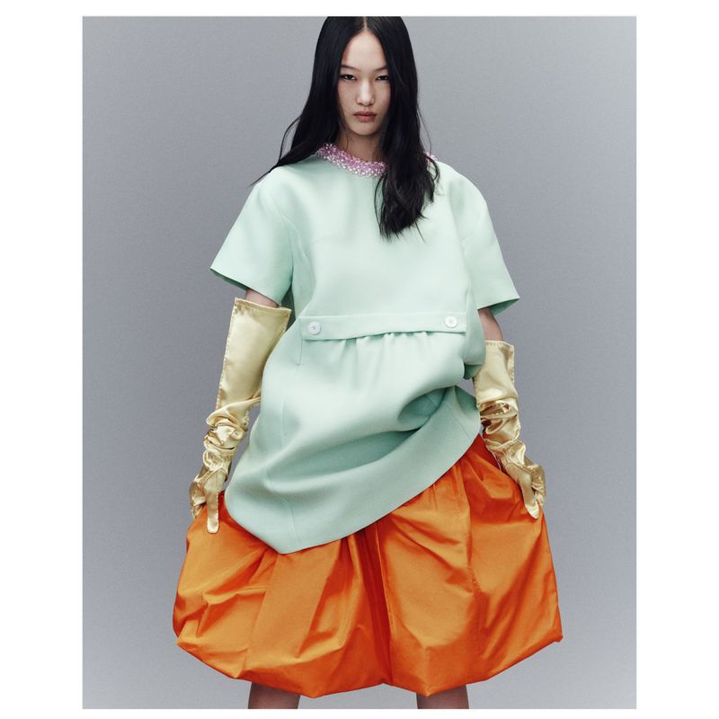 An Asian woman models in a light-green, short-sleeved dress with a pink-beaded collar and orange balloon skirt, in a fashion studio.