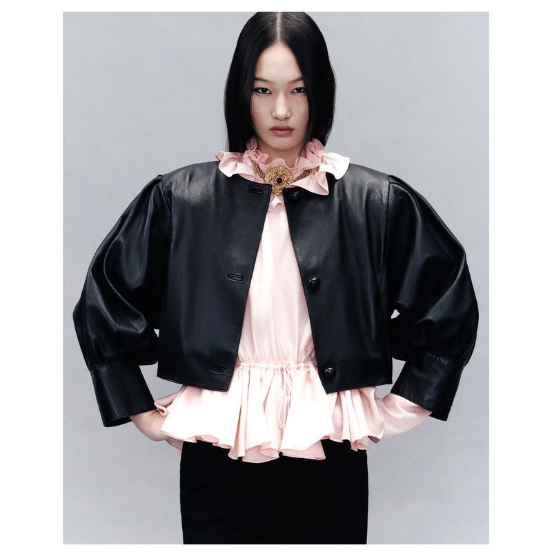 An Asian woman models a short black leather jacket with puffed sleeves, paired with a light pink blouse and black pencil skirt against a studio backdrop.