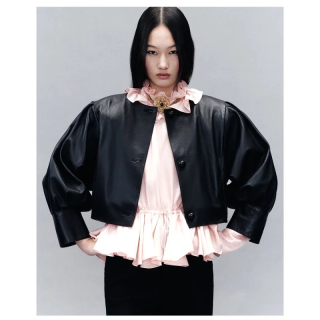 An Asian woman models a short black leather jacket with puffed sleeves, paired with a light pink blouse and black pencil skirt against a studio backdrop.