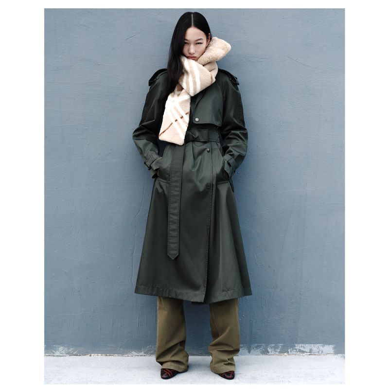 A stylish Asian woman in a green trench coat and patterned scarf poses against a gray wall in a full shot, looking elegant.