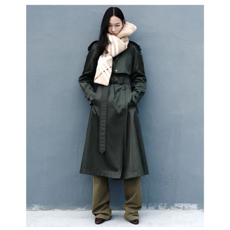 A stylish Asian woman in a green trench coat and patterned scarf poses against a gray wall in a full shot, looking elegant.