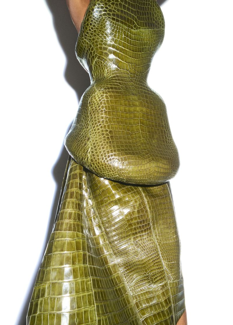 Close-up of a unique, green, crocodile-skin textured garment on a person, set against a bright white background, highlighting its abstract design.