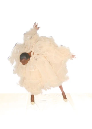 Model Debra Shaw wearing a voluminous beige tulle ruffle dress and matching pointed heels against a white studio background.