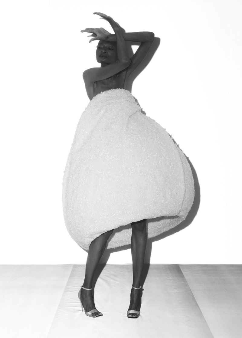 Full shot of a black woman in high heels, sculptural fashion skirt, and posed arms against a white background in a photo studio.
