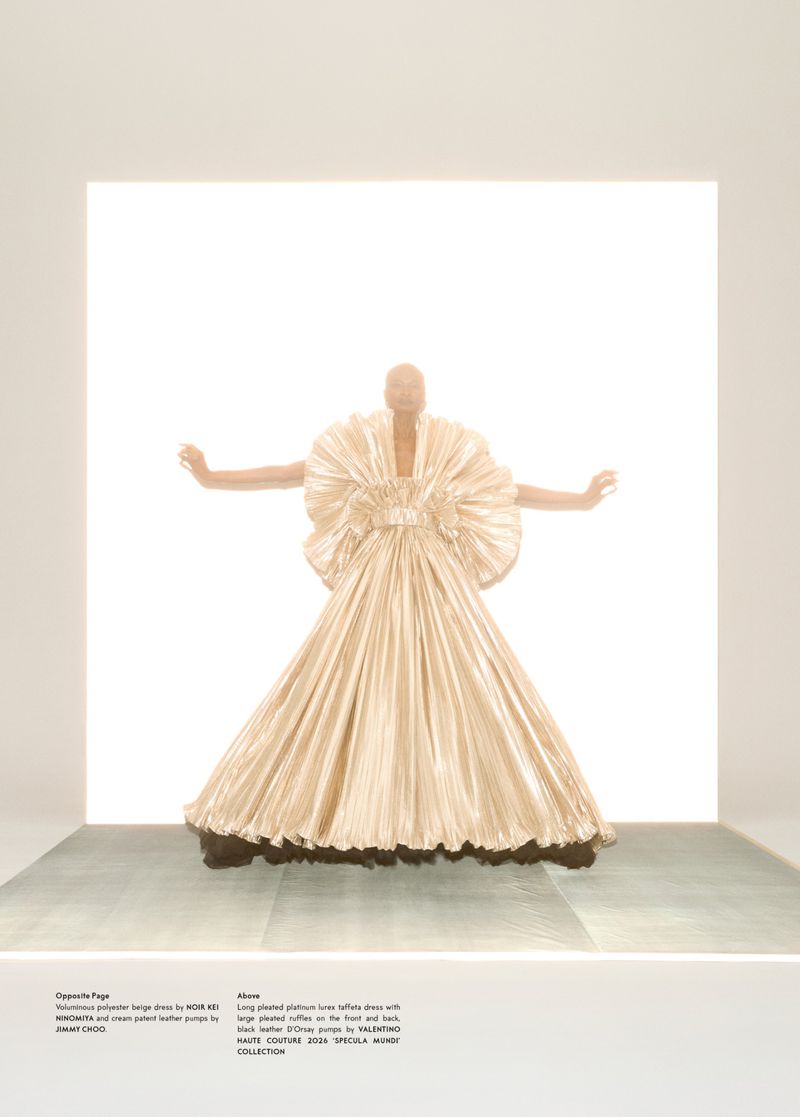 A lone model poses in a champagne-colored, floor-length couture dress, dramatically lit against a stark white backdrop in a fashion editorial.