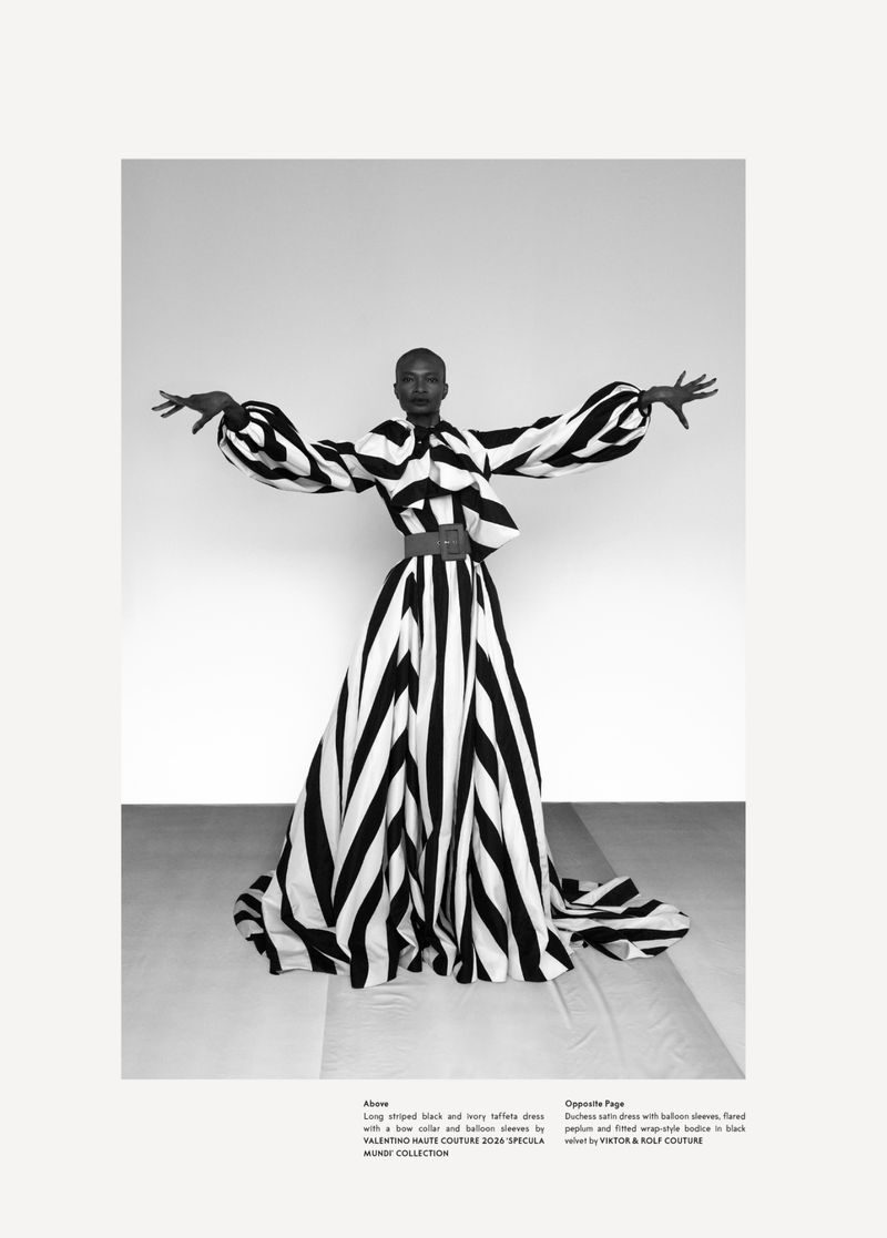 A black woman models a bold, black and white striped, avant-garde gown with balloon sleeves in a studio, striking a pose with arms outstretched.