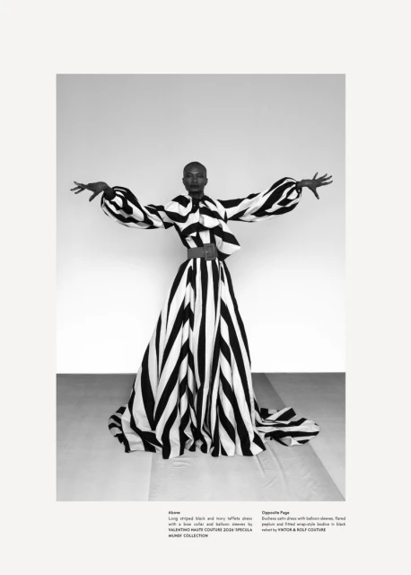 A black woman models a bold, black and white striped, avant-garde gown with balloon sleeves in a studio, striking a pose with arms outstretched.