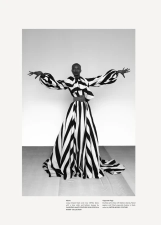 A black woman models a bold, black and white striped, avant-garde gown with balloon sleeves in a studio, striking a pose with arms outstretched.