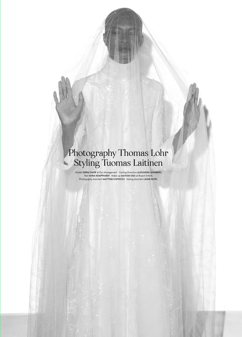 A model in an indoor photo studio setting, covered by a semi-transparent white veil, poses with her hands up in a fashion shot.