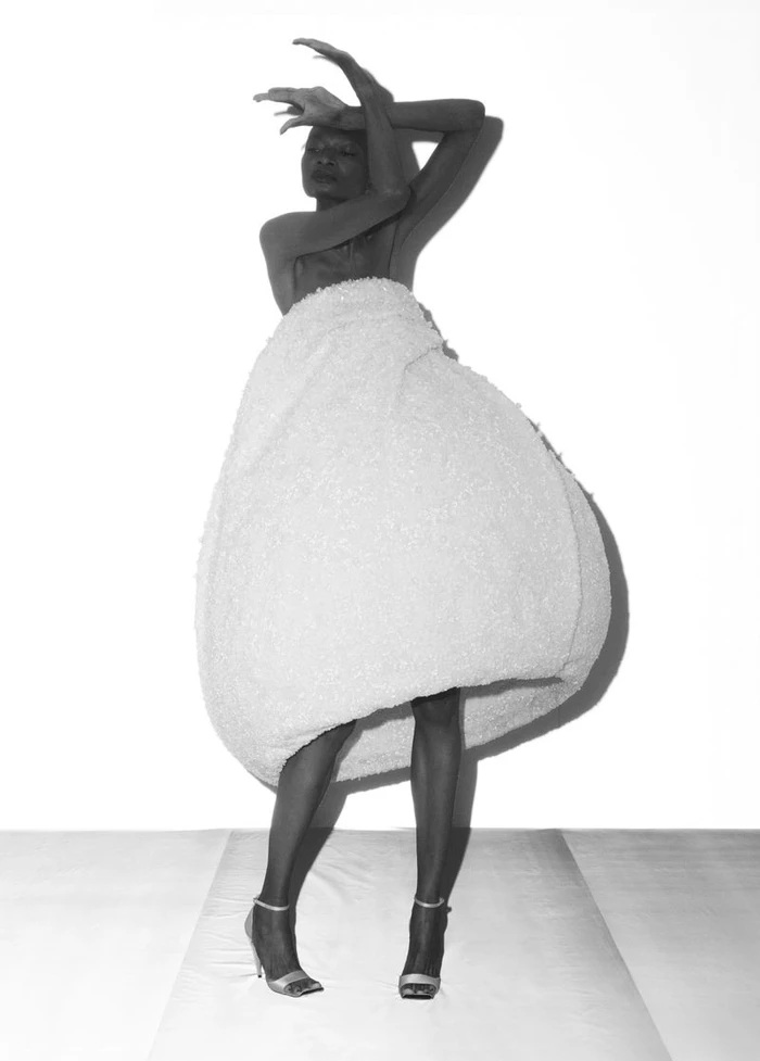 Model Debra Shaw posing in a voluminous sculptural white bulbous dress and high heels against a white studio backdrop.