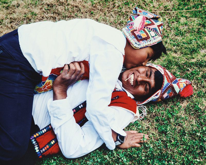 A couple, wearing colorful traditional hats, shares a happy moment lying in green grass. They wear white long sleeve shirts.