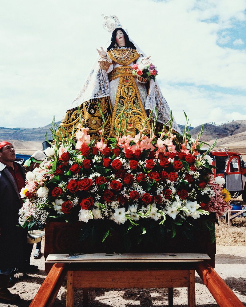 Statue of a religious figure adorned with flowers, carried by people outdoors against a backdrop of a clear sky and rolling hills.