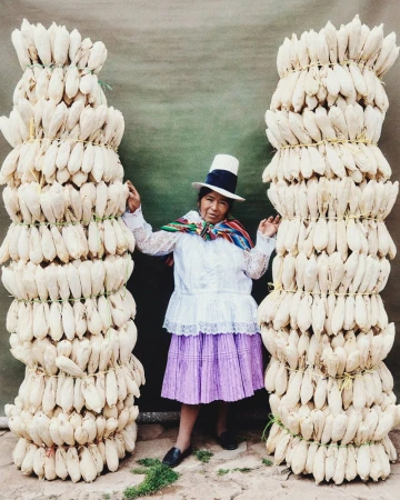 Portrait of indigenous Peruvian woman in traditional clothing standing between two stacks of corn. Cultural image featuring local produce.