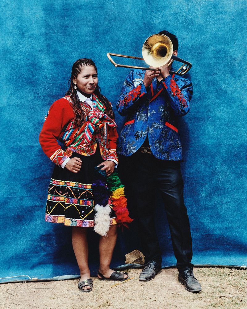 Two people in traditional clothing: a woman stands beside a man playing a trombone against a textured blue backdrop.