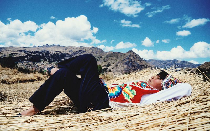 Person in traditional Andean clothing rests on a hay bed outdoors against a backdrop of mountains and a partly cloudy sky on a sunny day.