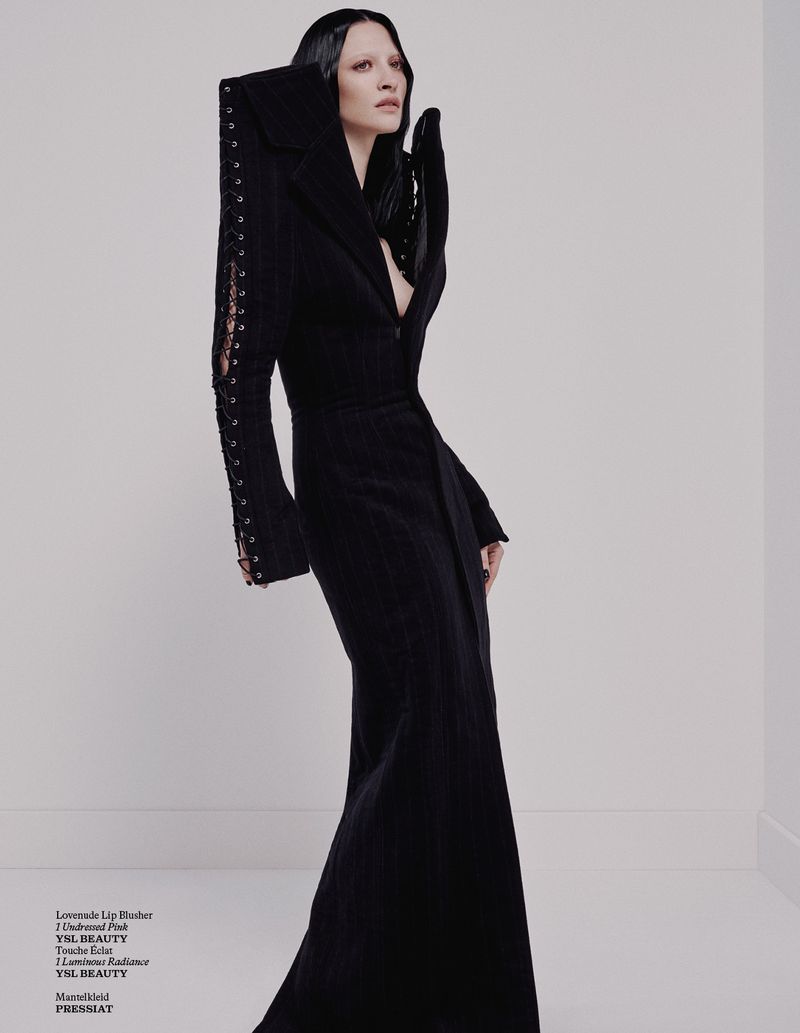 Woman poses in dramatic black gown with architectural shoulder details and sleeves against a neutral studio backdrop in a fashion editorial.