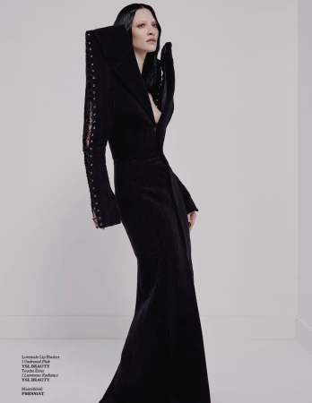 Woman poses in dramatic black gown with architectural shoulder details and sleeves against a neutral studio backdrop in a fashion editorial.