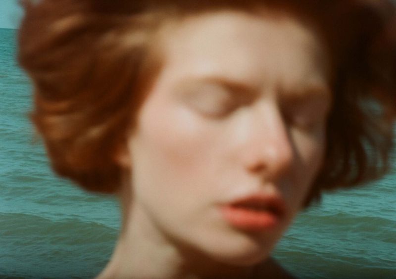 Close-up of a woman with auburn hair standing outside, facing downwards in a blurred composition with the ocean in the background.