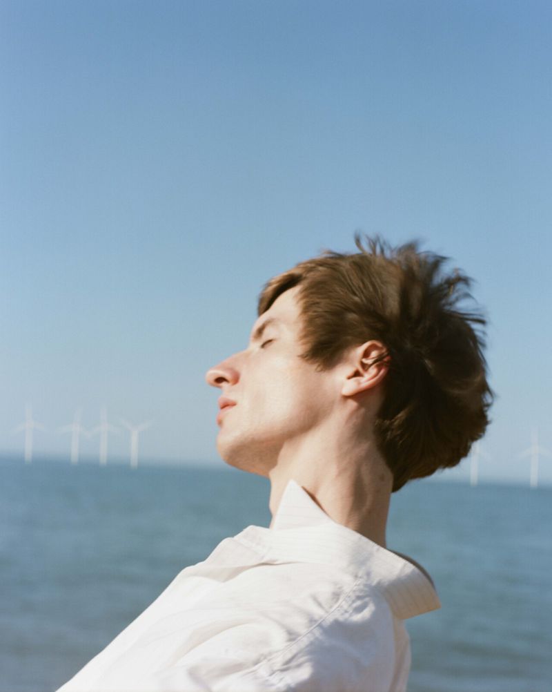 Man with eyes closed in white shirt stands near ocean on a sunny day with blue sky and wind turbines in the distance.
