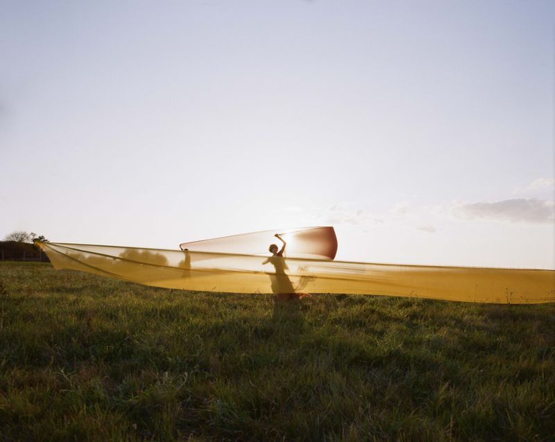 Woman in yellow dress holds translucent orange fabric in a grassy field at sunset, creating a flowing, ethereal visual.