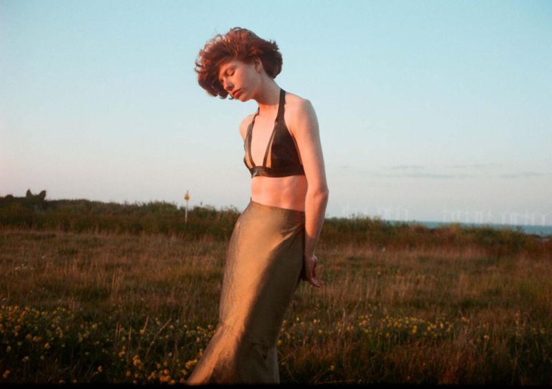 Adult female stands in a field wearing a skirt and bra top at sunset, with a grassy field and the sky in the background.