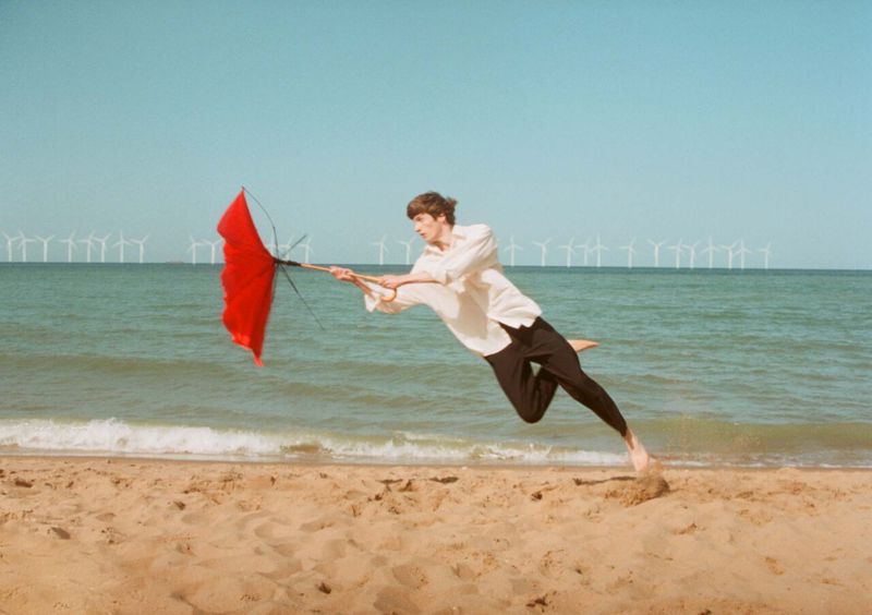 A young adult male is playfully airborne on a sandy beach, gripping a vibrant red umbrella against a backdrop of sea and wind turbines.