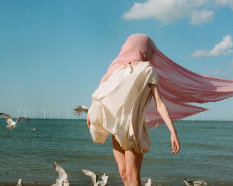 Woman in a flowing pink scarf and cream-colored dress standing on a beach, with seagulls and wind turbines in the background on a sunny day.