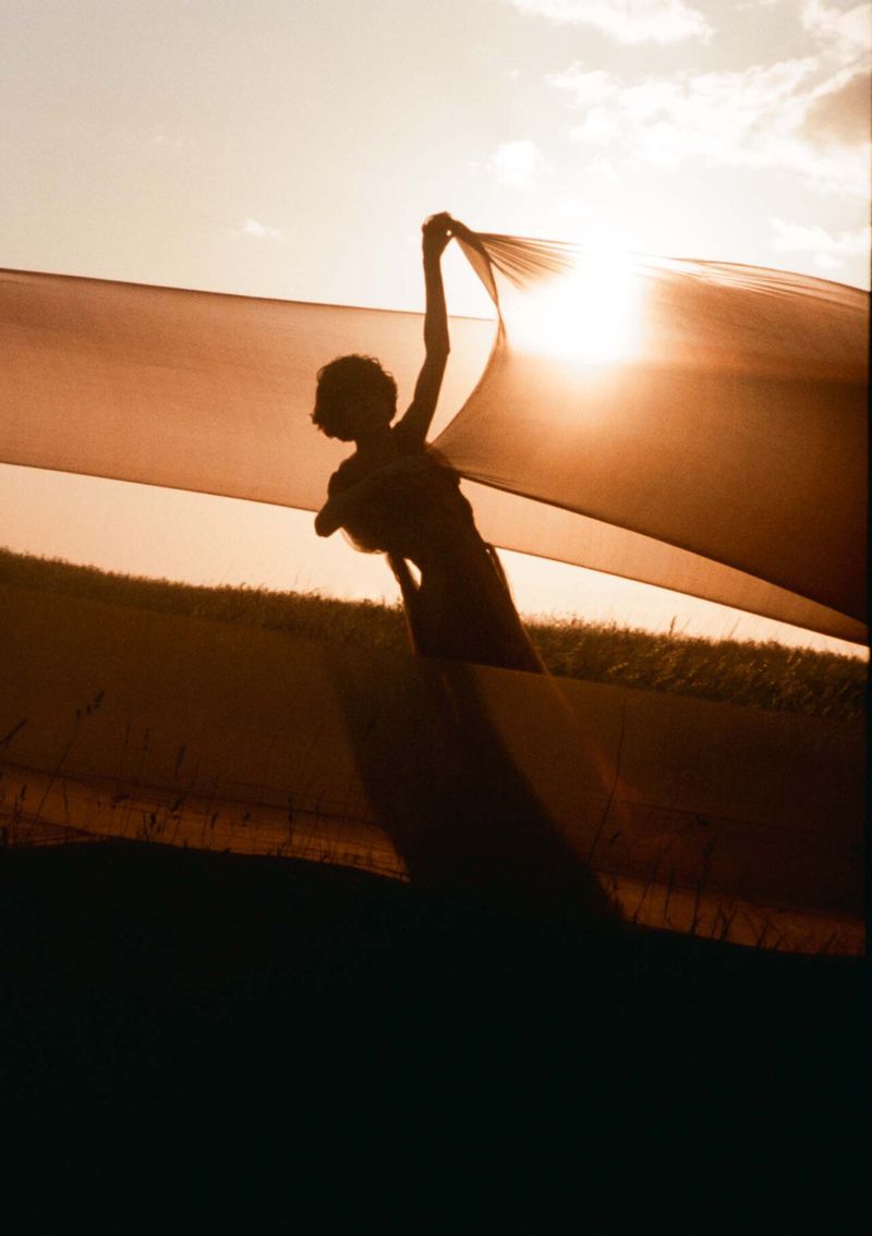 Silhouette of a woman holding a fabric against a bright sky at sunset.  Outdoor shot with a soft, warm color palette.